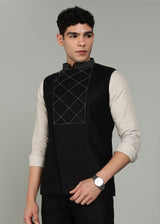 Man wearing a black embroidered 100% pure linen vest in 25 LEA over an ecru shirt on a gray background