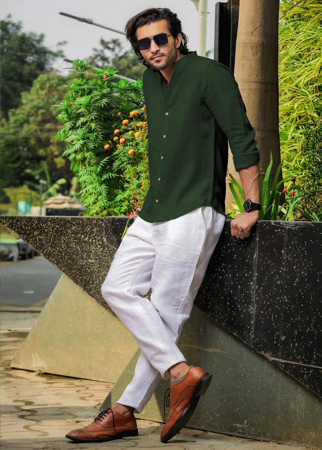 Man wearing a 100% pure linen dark fern green shirt, white pants, and brown shoes leaning against a wall with greenery in the background.