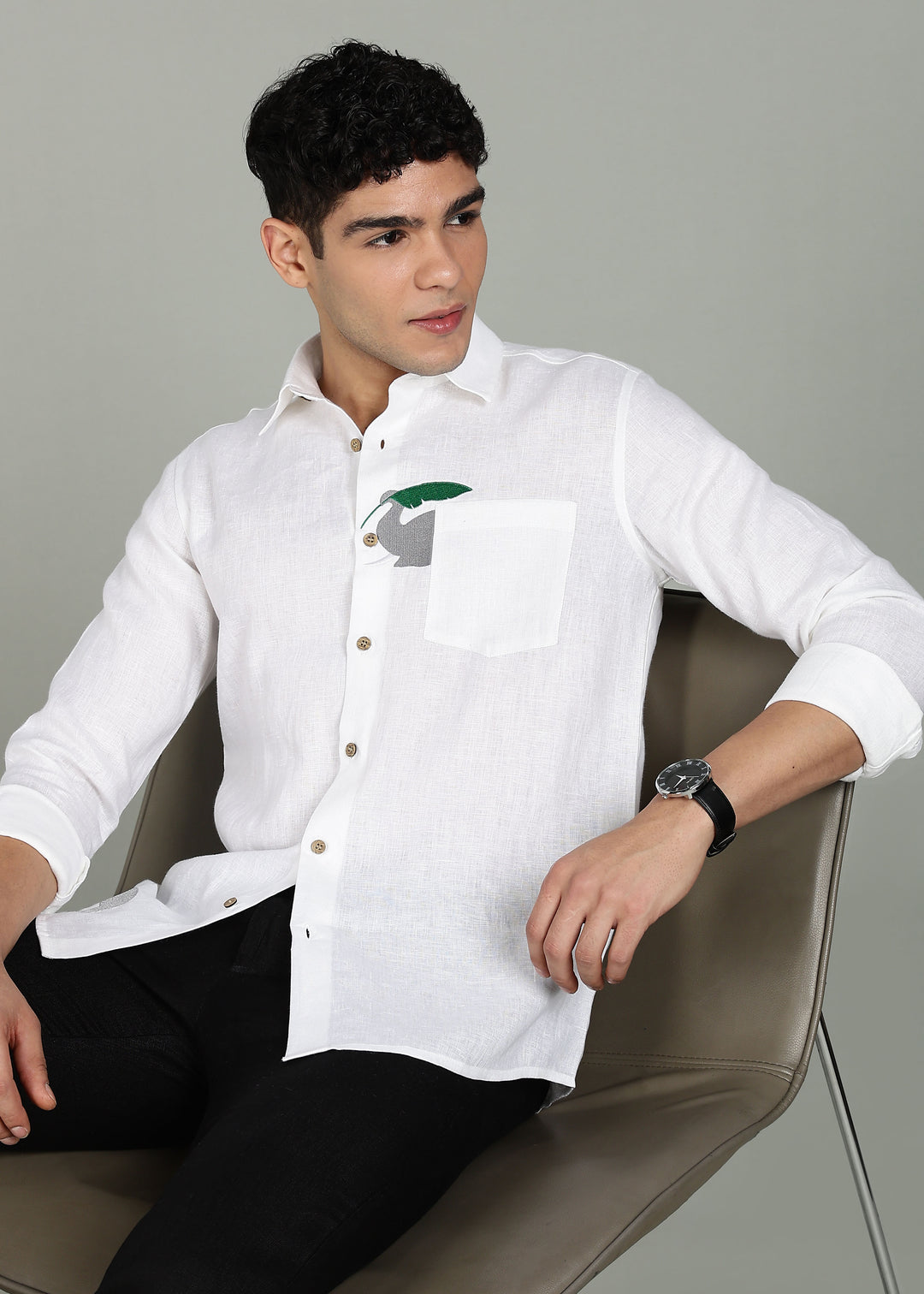 Man wearing a 100% pure linen white shirt in 44 LEA with elephant embroidery sitting on a chair against a gray background