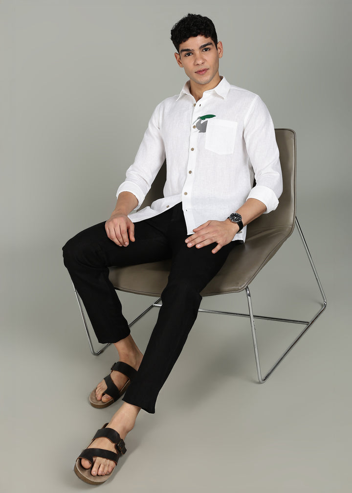 Man wearing a 100% pure linen white shirt in 44 LEA with elephant embroidery sitting on a chair against a gray background