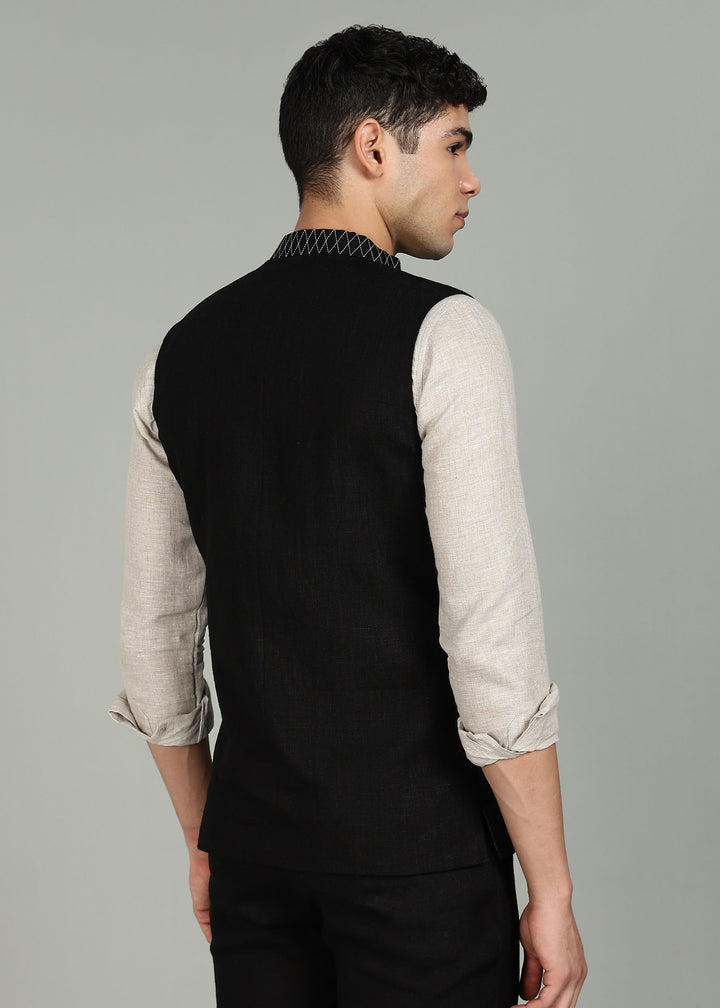 Man wearing a black embroidered 100% pure linen vest in 25 LEA over an ecru shirt on a gray background