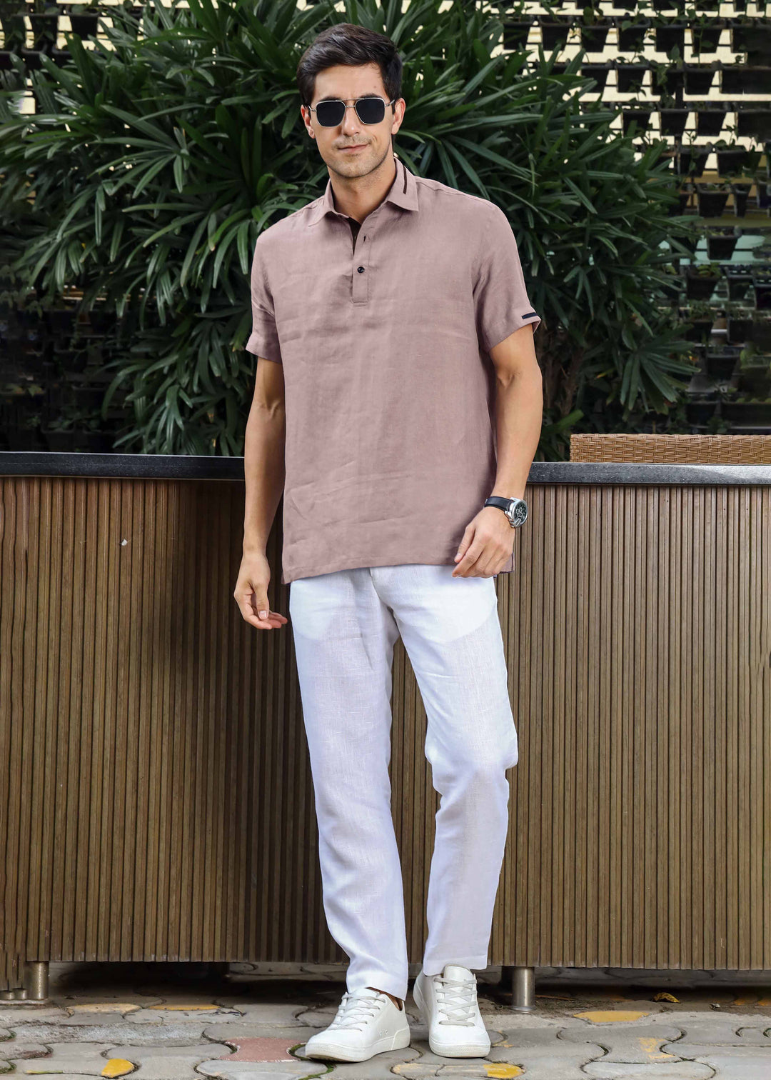 Man wearing a 100% pure linen cameo pink t-shirt and white pants standing outdoors with greenery in the background