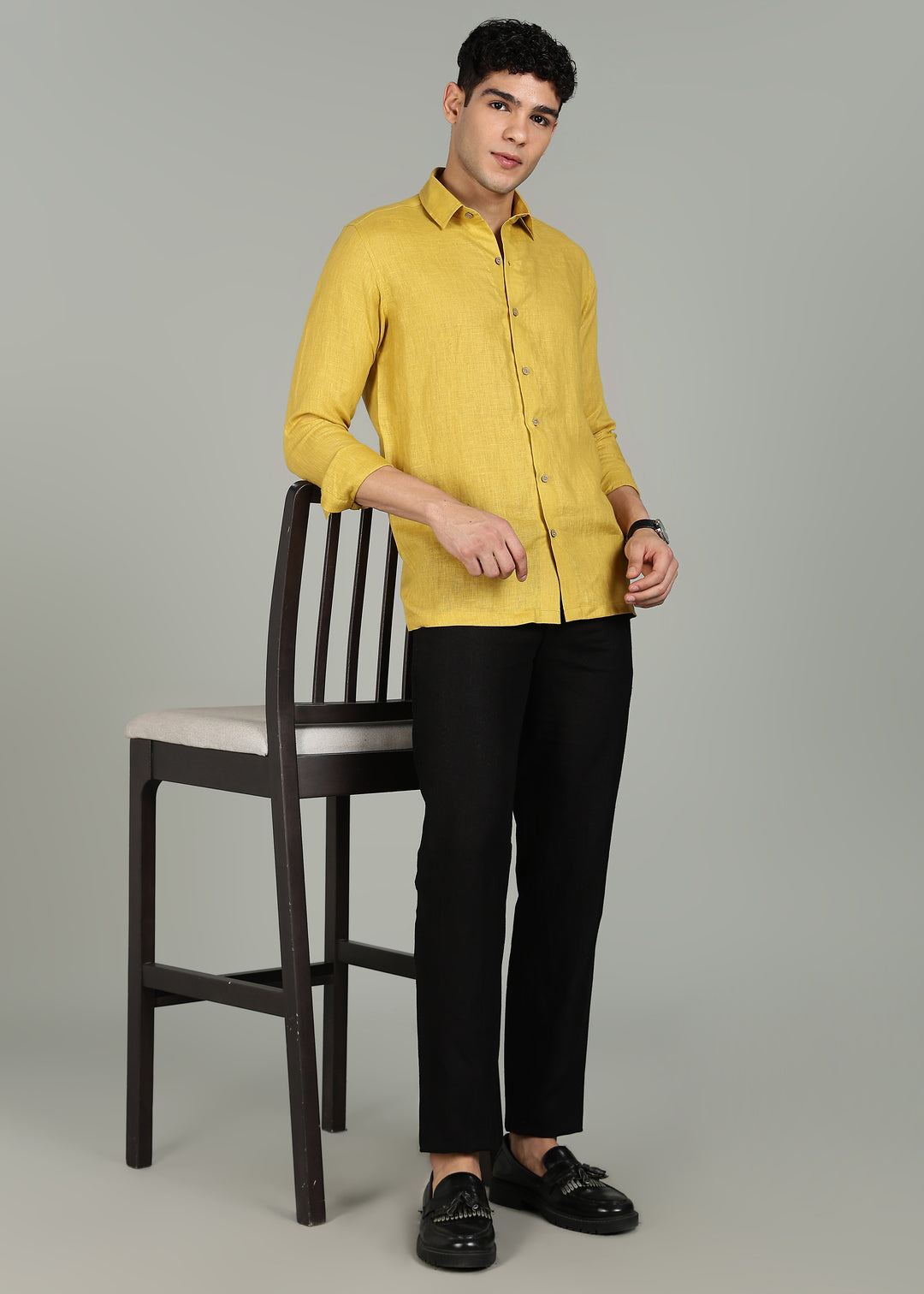 Man wearing a 100% pure linen sunburst yellow full sleeve shirt in 60 LEA and black pants standing next to a chair on a gray background