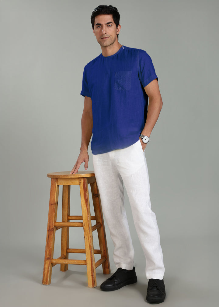 Man wearing a 100% pure linen egyptian blue t-shirt and white pants standing next to a wooden stool on a gray background