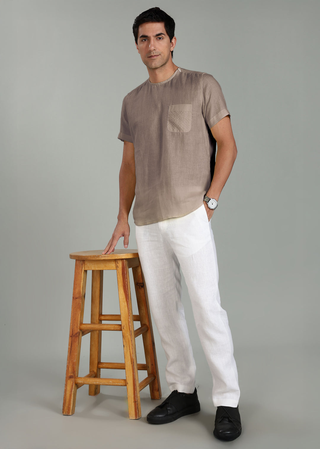 Man wearing a 100% pure linen fawn pink t-shirt standing next to a wooden stool on a gray background