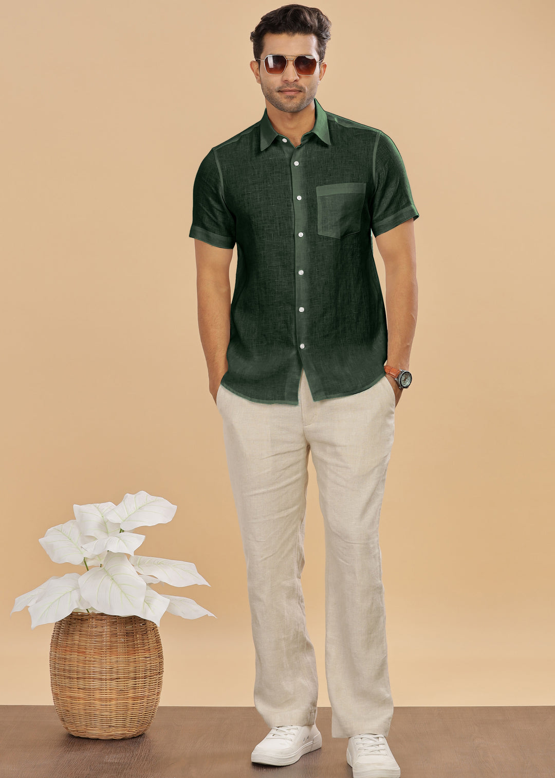 Man wearing a 100% pure linen dark fern green shirt and ecru pants standing next to a plant on a beige background