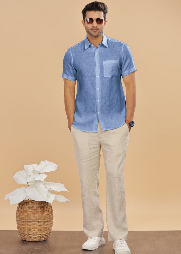 Man wearing a 100% pure linen olympic blue shirt and ecru pants standing next to a plant on a beige background